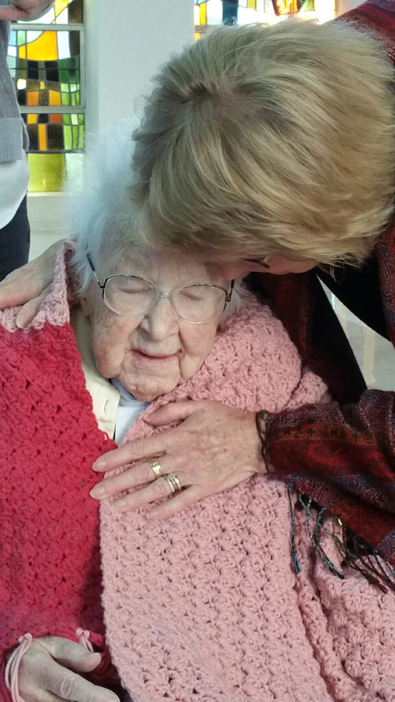 An elderly woman sits wrapped in an afghan with her eyes closed as another woman bends over to embrace her