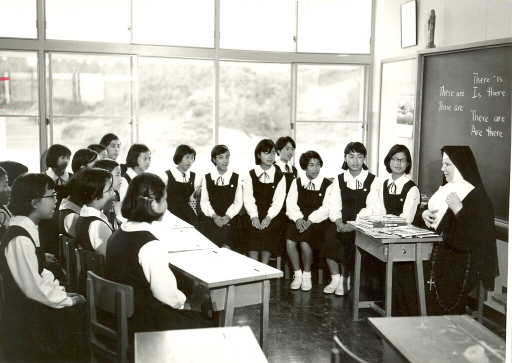 A black and white photo of a classroom with a dozen uniformed Japanese teenage girls gathered around a habited sister
