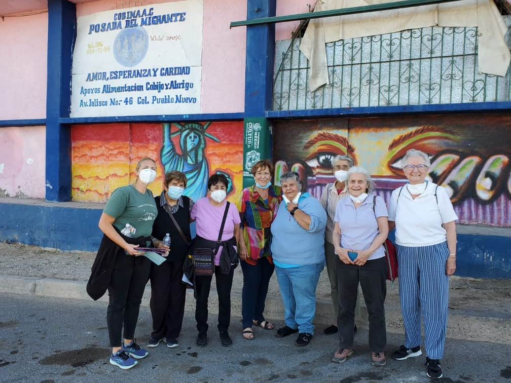 A group of sisters wearing masks pose in front of the sign for the shelter in Mexicali, Mexico and a large mural
