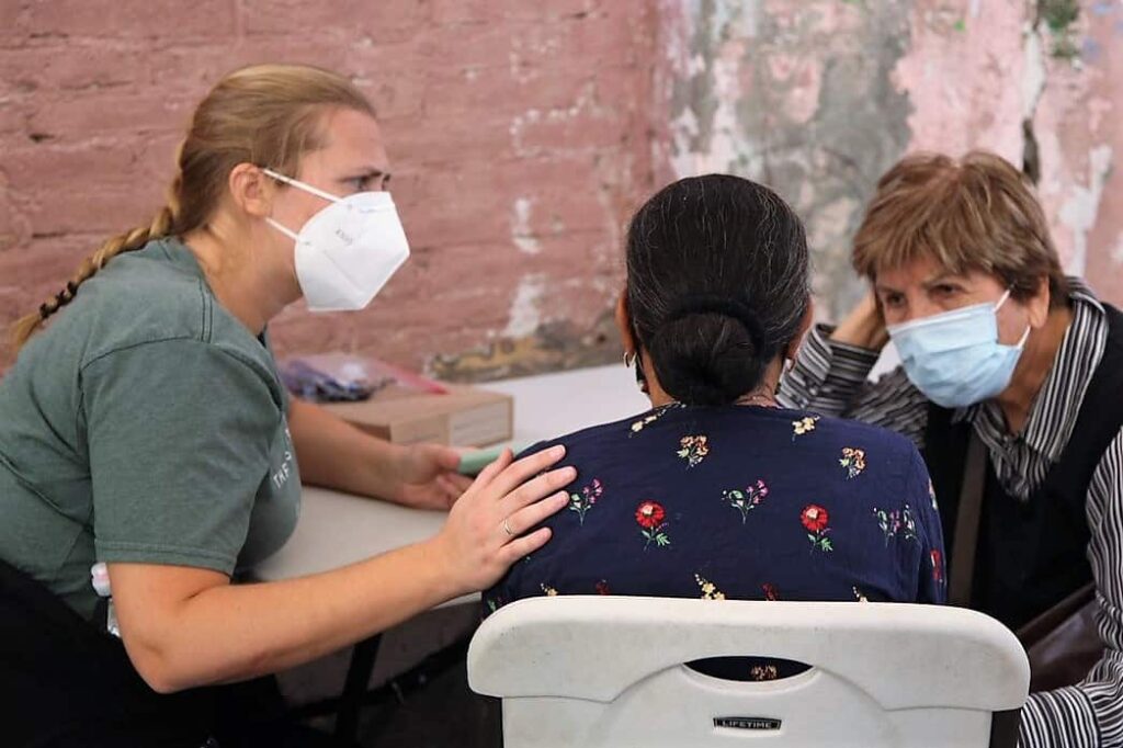 Two sisters, wearing masks, comfort a woman who has her back turned to the camera