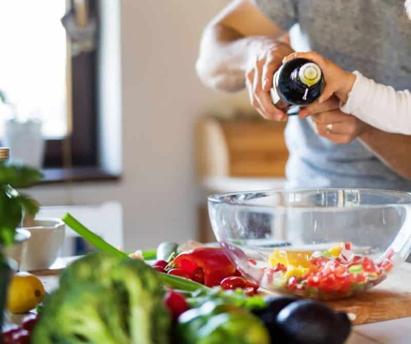 A child helps an adult pour oil onto a bowl of chopped vegetables