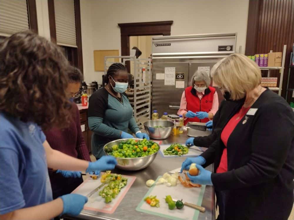 A group of five people stand around a commercial kitchen table prepping vegetables