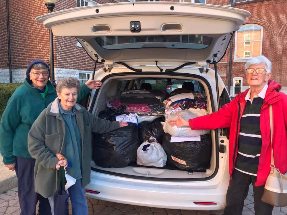 Three sisters gesture toward the trunk of an SUV, which is packed to the top with bags of donated items