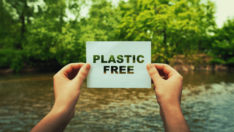 Two hands hold up a sign reading "Plastic free" in front of a pristine body of water surrounded by leafy trees