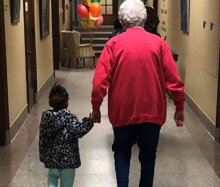 A small child holds the hand of an older sister as they walk down the hall. Colorful balloons float in the distance.