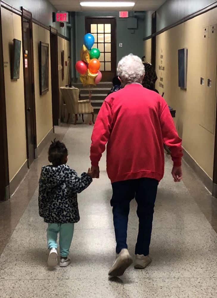 A small child holds the hand of an older sister as they walk down the hall. Colorful balloons float in the distance.