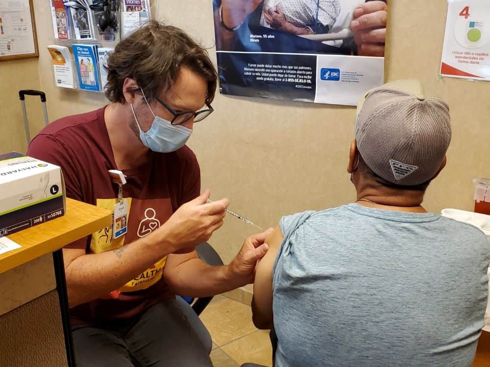 A man wearing a mask administers a shot in the arm of a man with his back to the camera