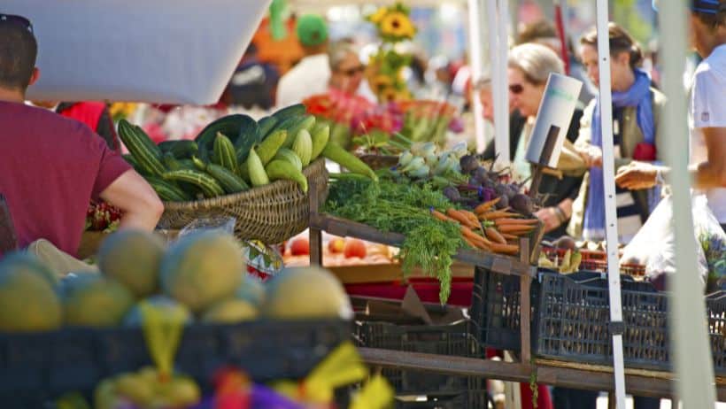 Produce is piled high on tables at a farmers market and shoppers browse