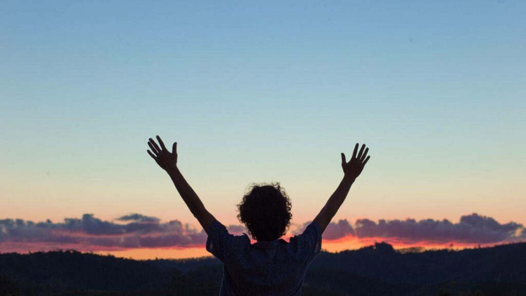 a woman in silhouette raises her hands above her head in praise for the a beautiful sunset in the background