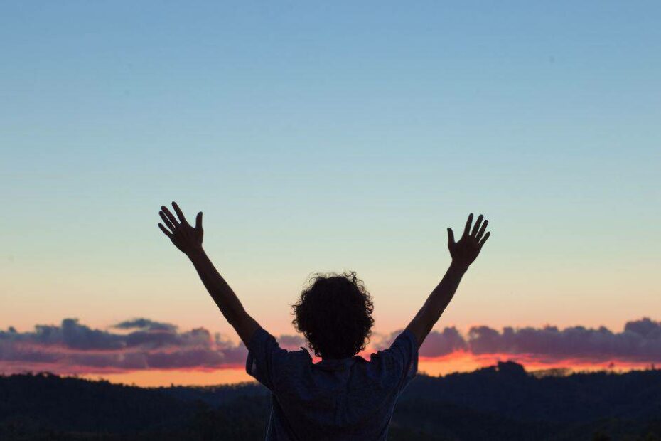 a woman in silhouette raises her hands above her head in praise for the a beautiful sunset in the background