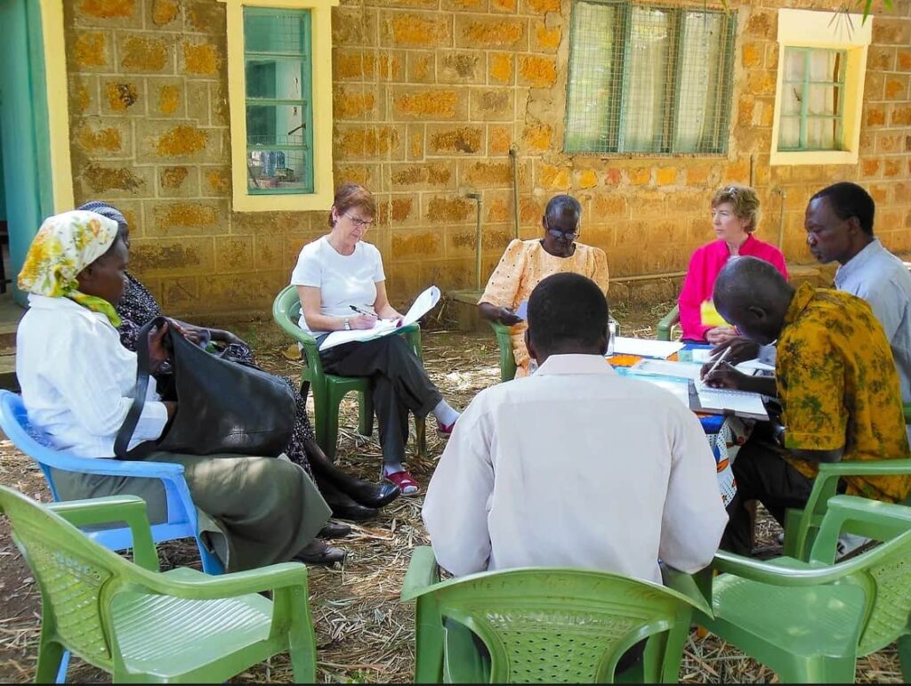 a group of people sits outside in a circle of green plastic chairs. One person is writing in an open notebook