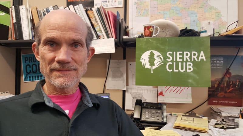 John Hickey sits in front of a work desk and bookshelves. A Sierra Club sign hangs over his shoulder