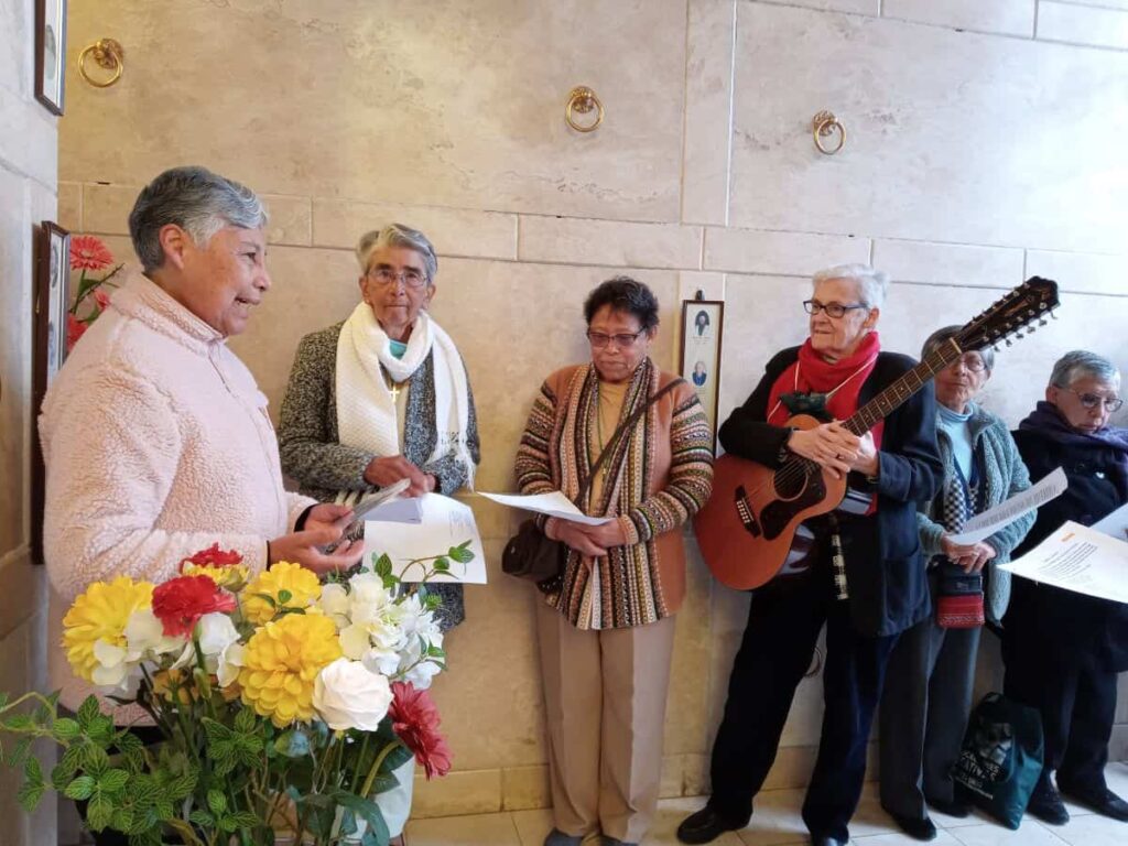 Six sisters gather in a mausoleum with flowers in the foreground. One holds a guitar and the others hold programs 