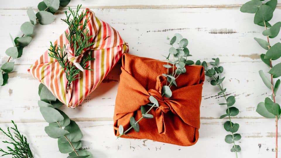 Two gifts wrapped in red cloth and eucalyptus leaves sit on a light wood table