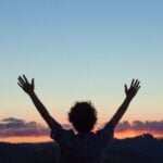a woman in silhouette raises her hands above her head in praise for the a beautiful sunset in the background