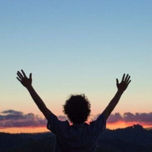 a woman in silhouette raises her hands above her head in praise for the a beautiful sunset in the background