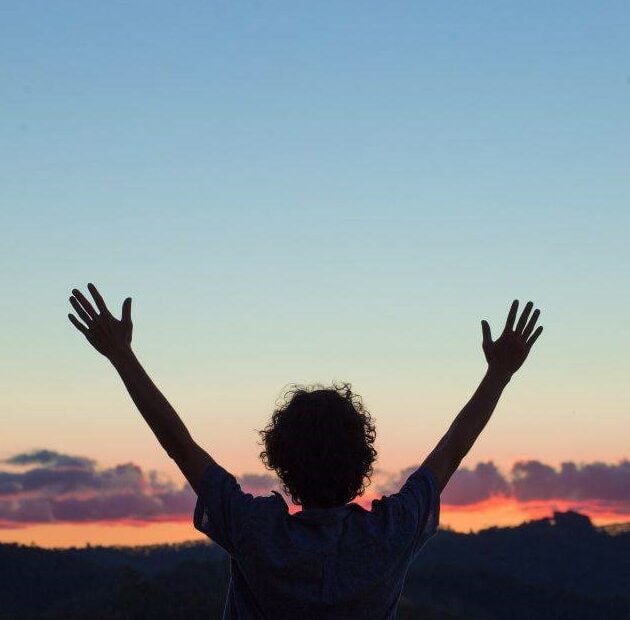 a woman in silhouette raises her hands above her head in praise for the a beautiful sunset in the background