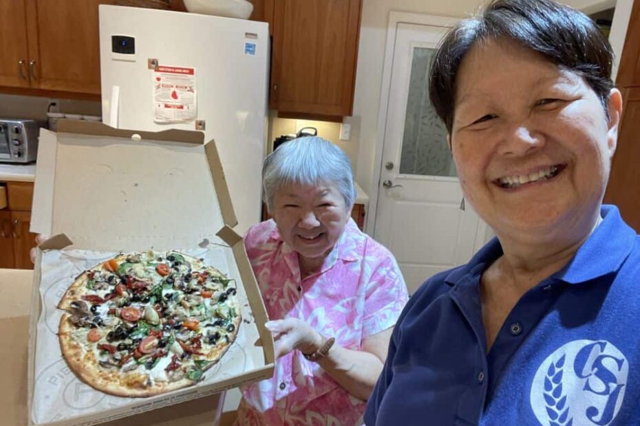 Two sisters smile broadly in their kitchen. One is holding open a pizza box with a vegetarian pizza
