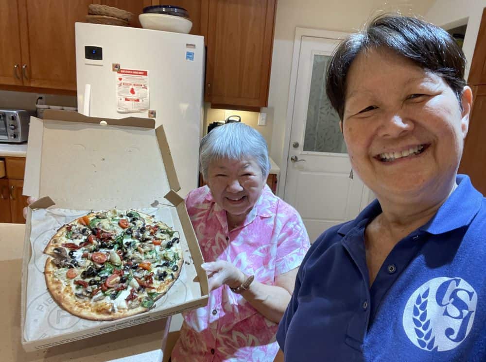 Two sisters smile broadly in their kitchen. One is holding open a pizza box with a vegetarian pizza