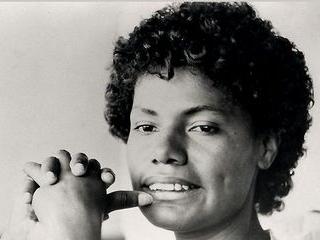 A black and white picture of María Elena Moyano, a young woman with short curly hair. We see her from the neck up. Her eyes are looking downward, and her clasped hands are near her face, with her thumb resting on her bottom lip.