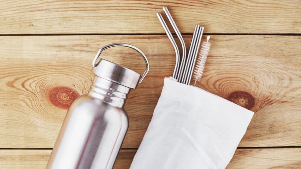 a metal water bottle and a cotton bag with metal reusable straws sticking out of it are laid out on an unfinished wood background