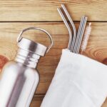 a metal water bottle and a cotton bag with metal reusable straws sticking out of it are laid out on an unfinished wood background