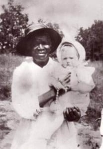 A grainy sepia-toned photo shows an older Black woman in 19th century attire holding a white baby in a white gown and bonnet. They are pictured outside.