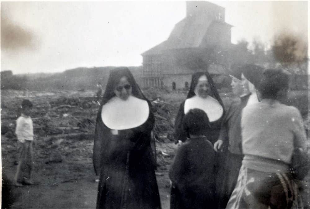 An old black and white photo shows four sisters wearing habits speaking a child and a woman. A mess of tsunami rubble extends to the horizon.