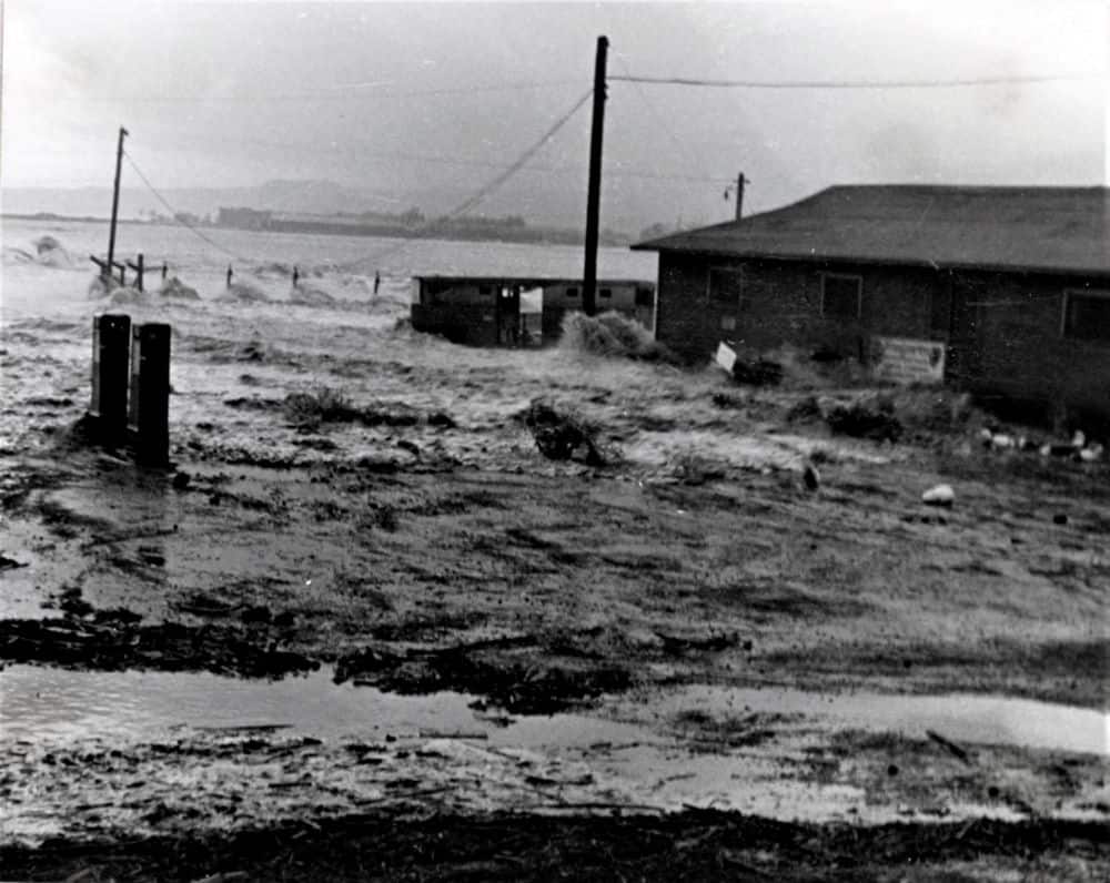 An old black and white photograph depicts a one story structure in the background, surrounded by water, mud and debris. A powerline sags dangerously low.