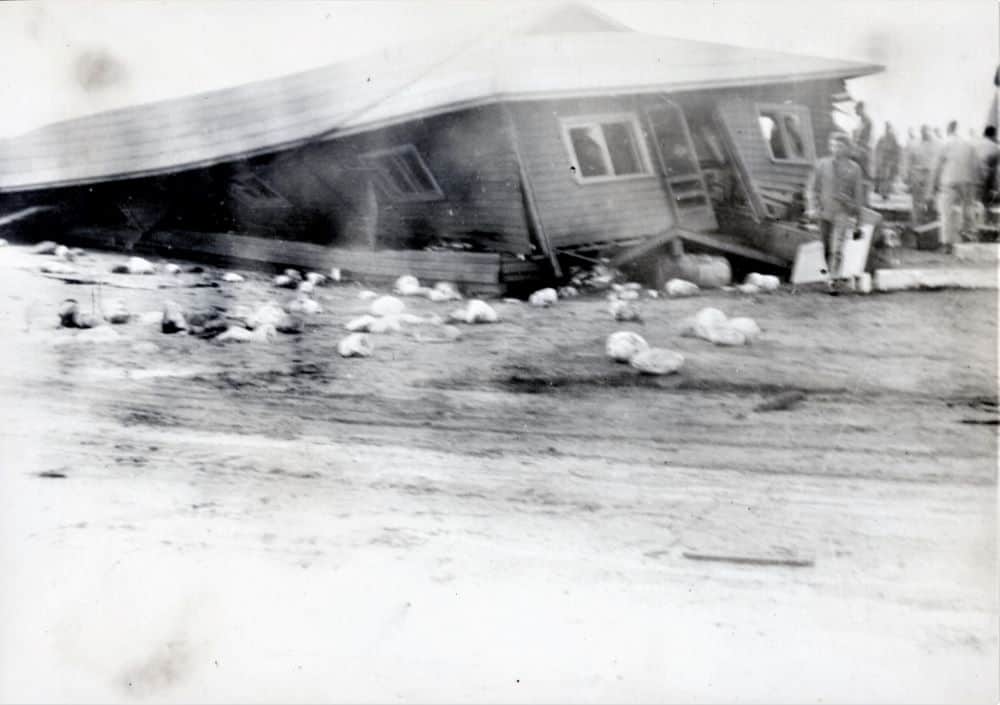 A blurry black and white photo depicts a one-story structure that has been shifted and crushed. A line of people are walking around the backside of the building. Detritus is scattered in the foreground.
