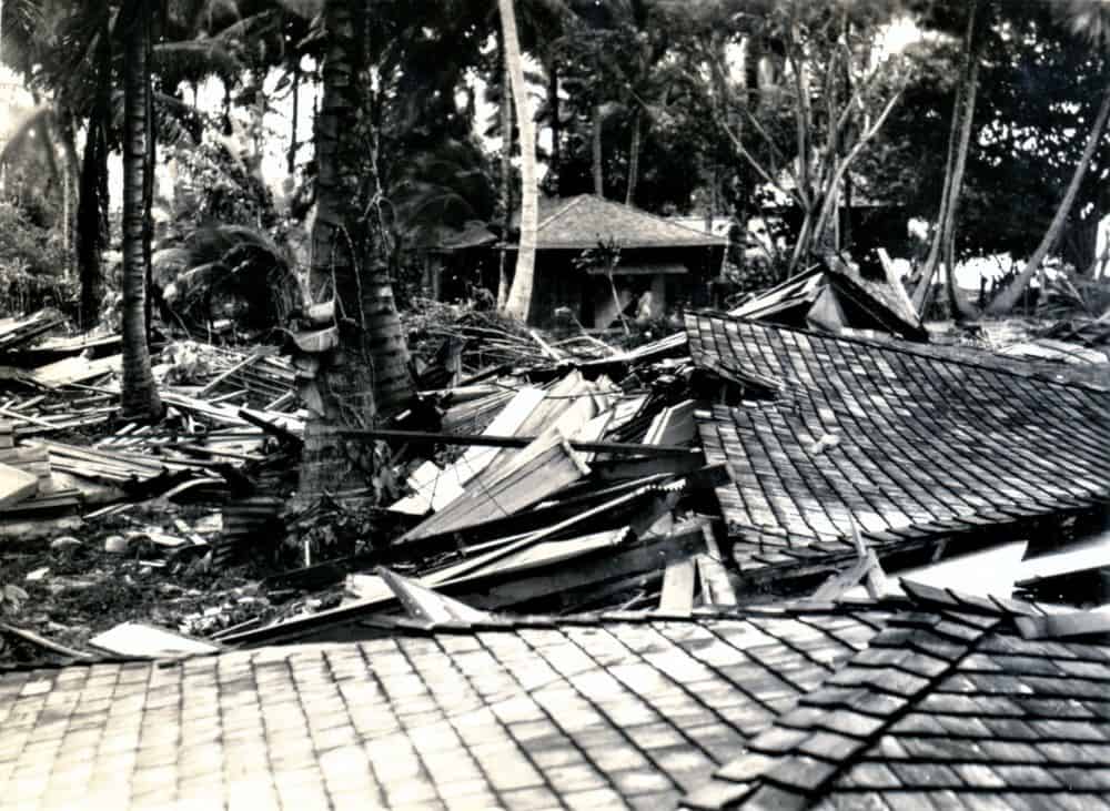 A black and white photo of flattened structures. Boards and collapsed roofs are scattered among palm trees.