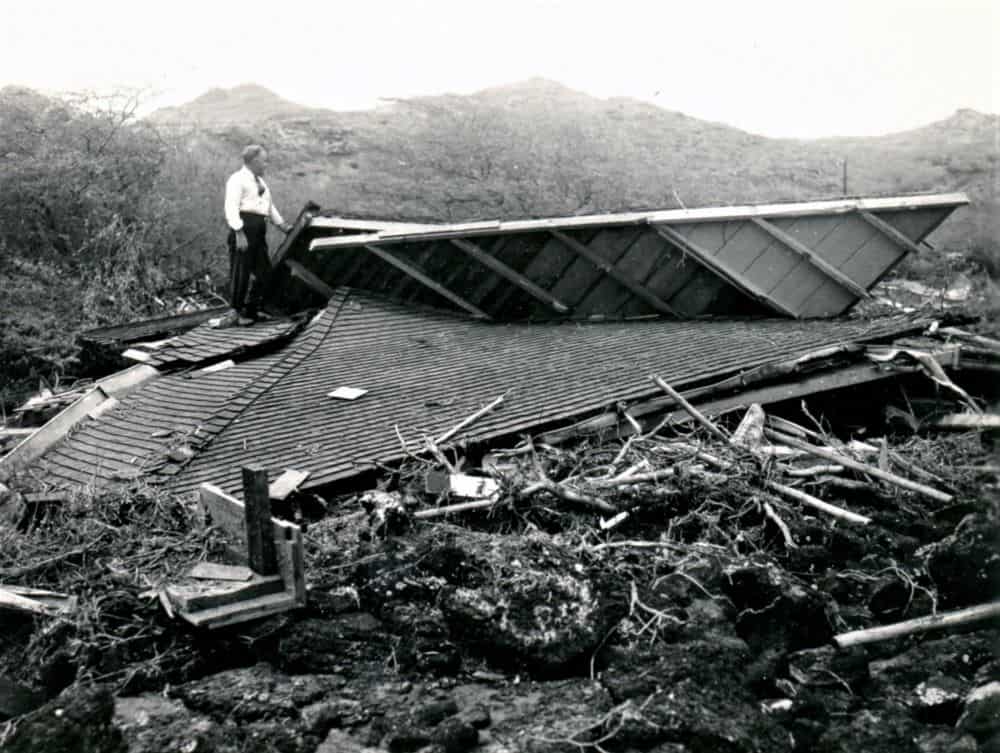 Black and white photo of a man in a white shirt and black pants standing on the flat roof of a collapsed building. Tree roots, sticks and mud are in the foreground. Mountains are visible on the horizon.
