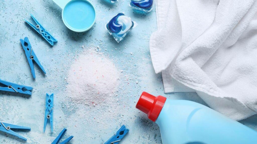 A haphazard scattering of blue plastic clothespins, a blue bottle of liquid laundry detergent with a red cap, laundry detergent pods and a pile of powdered detergent surround a white towel.