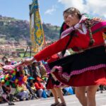 A young indigenous woman in traditional Peruvian dress spins while dancing in front of a crowd celebrating in the streets