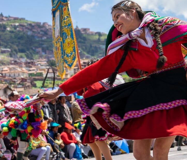 A young indigenous woman in traditional Peruvian dress spins while dancing in front of a crowd celebrating in the streets