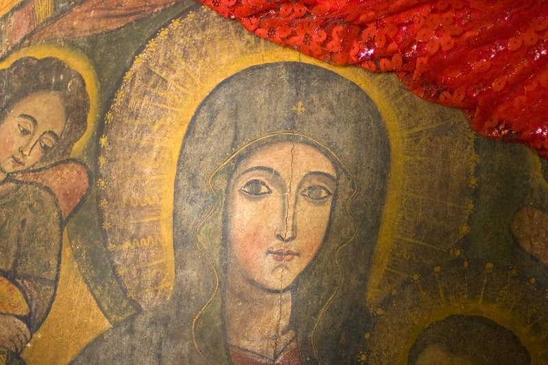 A photo of a fresco depicting the Virgin Mary wearing a veil and staring directly at the viewer. An angel appears over her shoulder