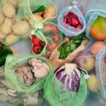 A colorful array of produce sits on a gray countertop in green and white reusable mesh produce bags