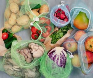 A colorful array of produce sits on a gray countertop in green and white reusable mesh produce bags