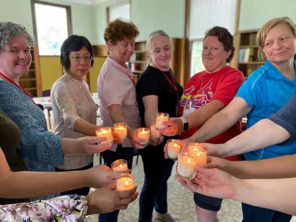 A group of 10 women stand in a circle extending their arms holding lit votive candles into the center