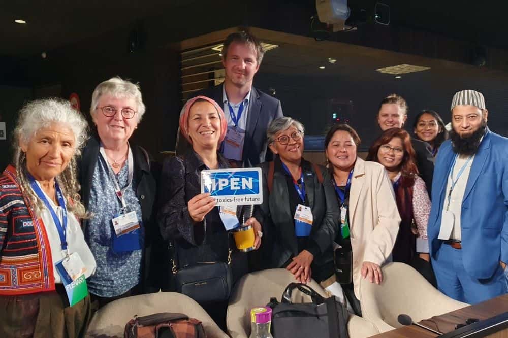 A multi-ethnic group of 10 people stands together behind a desk in a darkened room. A woman with a headscarf in the middle holds a sign reading "IPEN."