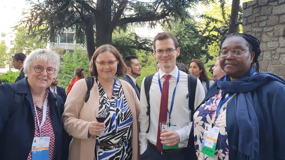 Four people stand together at an outdoor reception. They are wearing lanyards and holding glasses of wine.