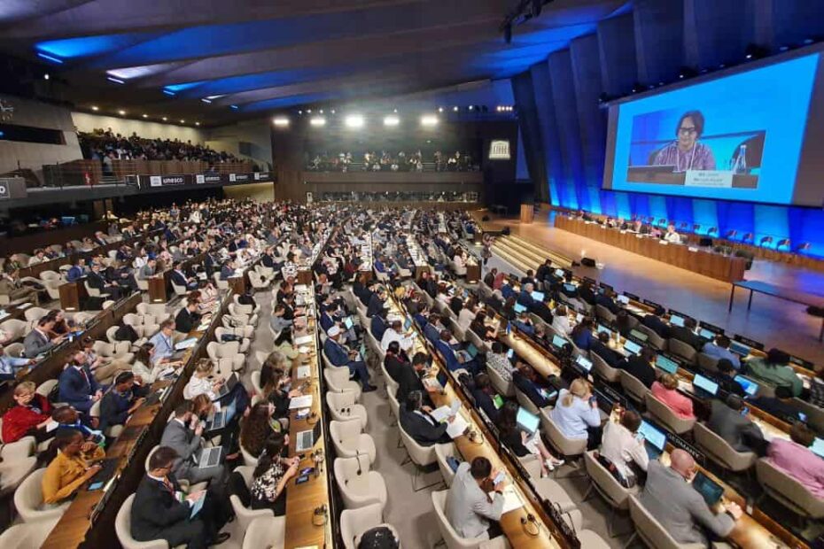 A huge room full of people sitting in rows of chairs and counters. Six people sit at a long table on the stage facing the crowd. One speaker appears on a giant screen at the front.