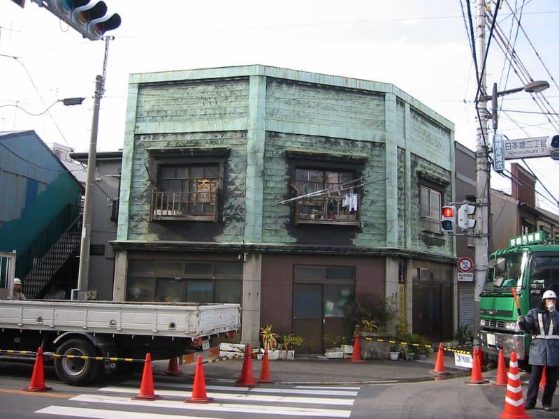 An urban grungy two-story turquoise building stands on a streetcorner, surrounded by orange cones and trucks. A helmeted worker directs traffic