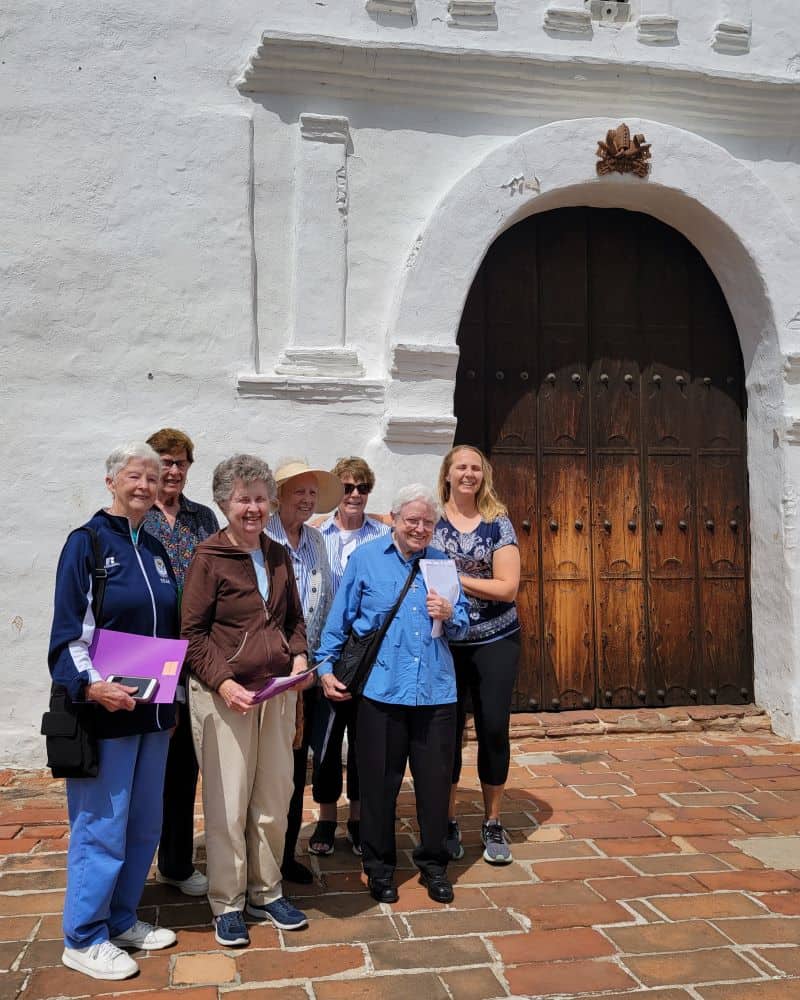 A color photo of seven women standing in front of a large wooden door in an adobe structure in 2023