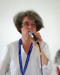 Sister Nathalie Becquart speaks into a microphone. She is wearing a white tunic shirt and a blue lanyard with white lettering on it. She stands in front of a white wall.