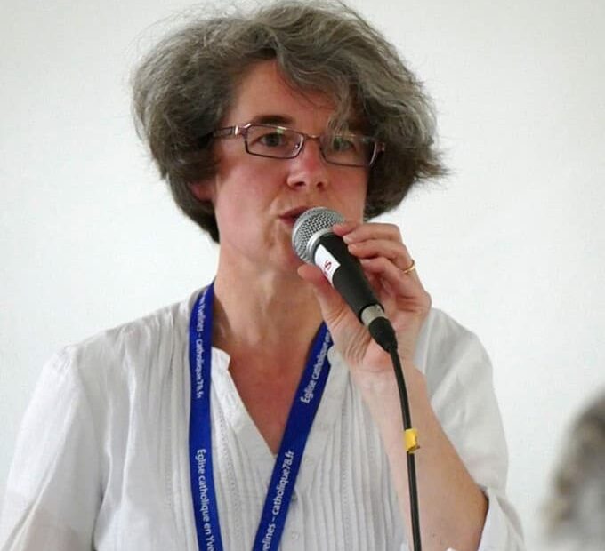 Sister Nathalie Becquart speaks into a microphone. She is wearing a white tunic shirt and a blue lanyard with white lettering on it. She stands in front of a white wall.