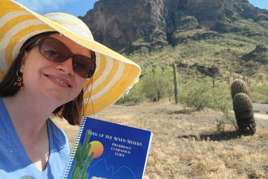 Woman wearing a blue tshirt, sunglasses and a wide brimmed yellow and white striped hat stands in front of a mountain in the desert sun. She is holding a book titled "The Trek of the Seven Sisters."