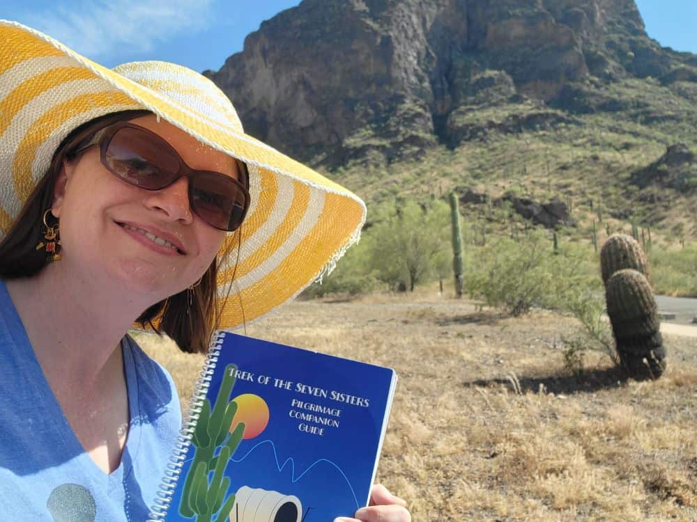 Woman wearing a blue tshirt, sunglasses and a wide brimmed yellow and white striped hat stands in front of a mountain in the desert sun. She is holding a book titled "The Trek of the Seven Sisters."