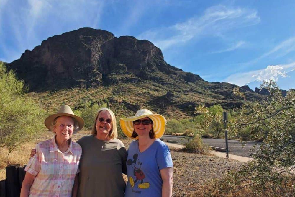 Thee women in straw hats and sunglasses stand together in front of a mountain in the desert.
