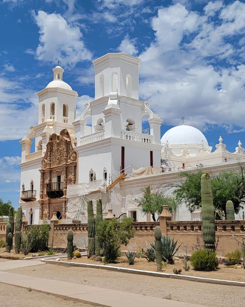 A bright white mission church stands against a blue sky surrounded by cacti